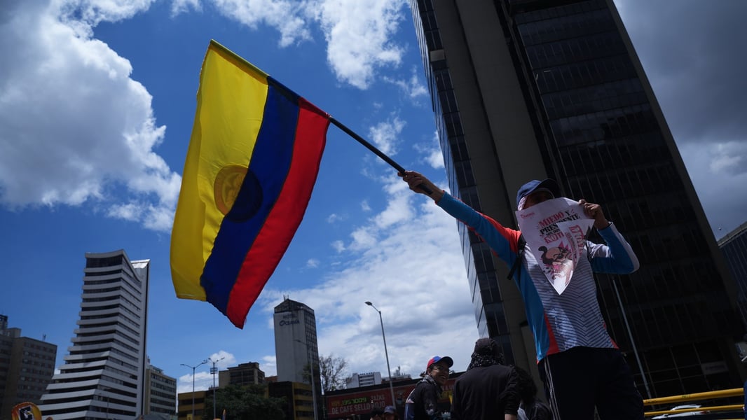 A protester in a blue cap holds up a Colombian flag and a placard against a Bogota skyline