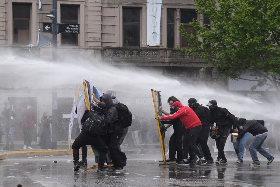 Hooded protesters crouch behind makeshift shields as a water cannon sprays them on a Buenos Aires street