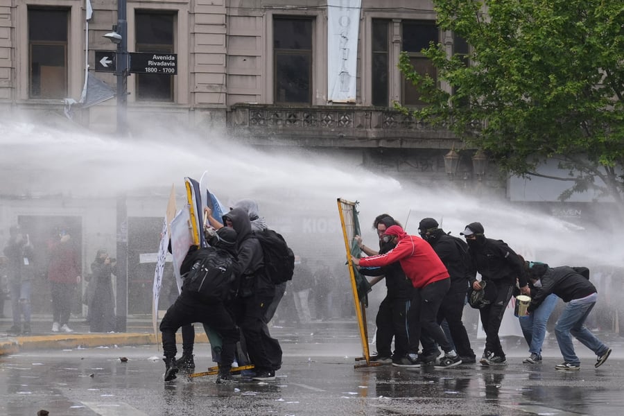 Hooded protesters crouch behind makeshift shields as a water cannon sprays them on a Buenos Aires street