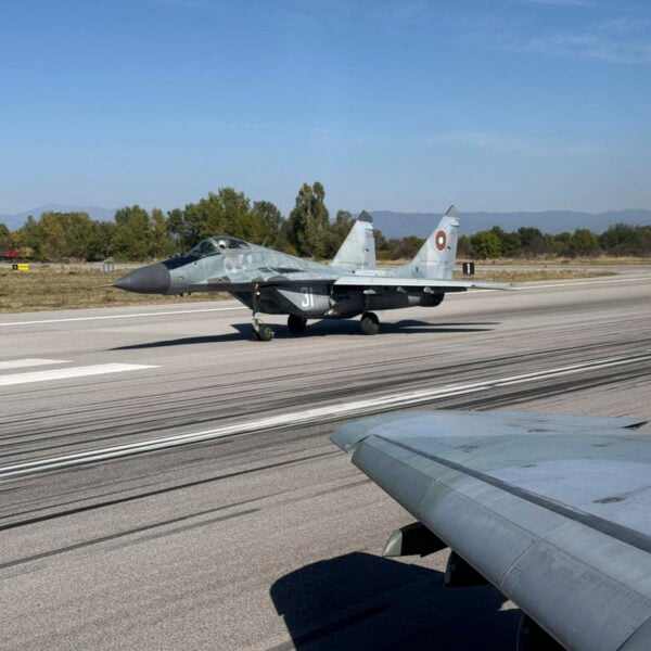 A grey Bulgarian Air Force MiG-29 fighter taxiing on a runway beneath a clear sky, seen past the wing of another aircraft
