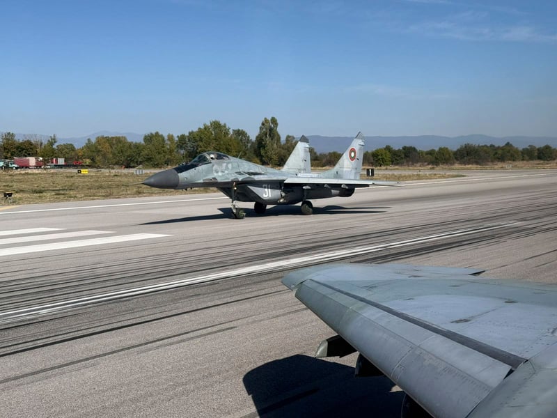 A grey Bulgarian Air Force MiG-29 fighter taxiing on a runway beneath a clear sky, seen past the wing of another aircraft