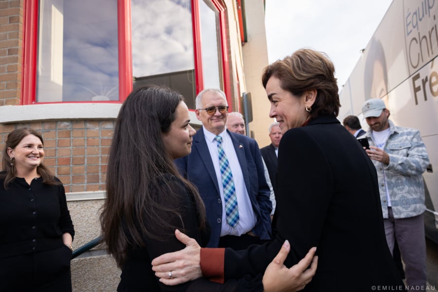 Two women embrace outside a brick building beside a CAQ campaign tour bus as supporters look on