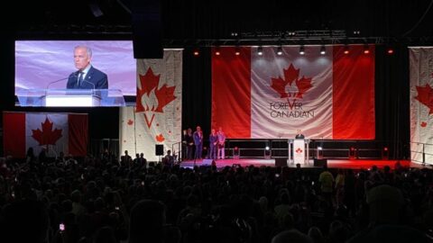 Prime Minister Mark Carney speaks at a podium in front of a Forever Canadian banner at an Edmonton rally, with his image on a large screen at left