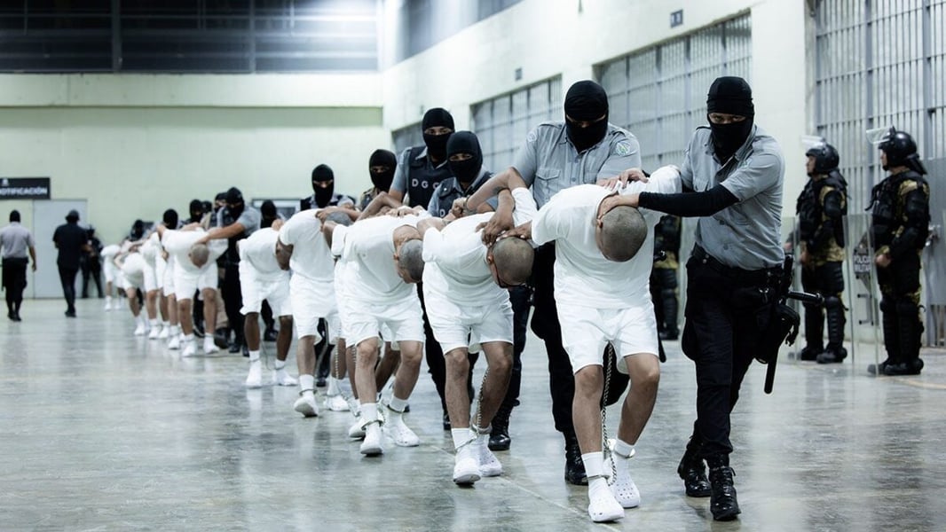 Masked Salvadoran guards escort a line of shirtless detainees in white shorts through the CECOT prison
