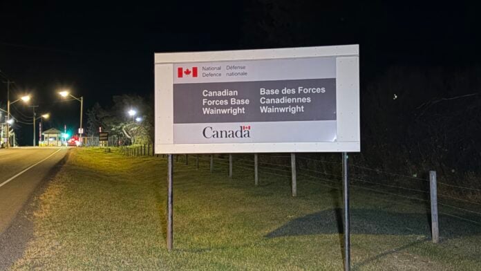 Sign reading Canadian Forces Base Wainwright beside a road at night, with the base gate and street lights in the distance