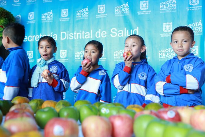 Young children in blue school tracksuits hold and bite into apples in front of a Bogota health department backdrop, with fruit in the foreground