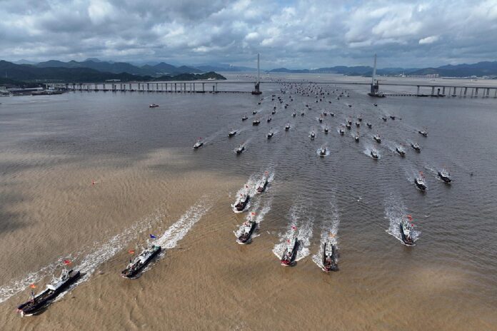 Aerial view of dozens of fishing boats with red flags speeding in a wedge formation past a long bridge as they leave a Chinese port
