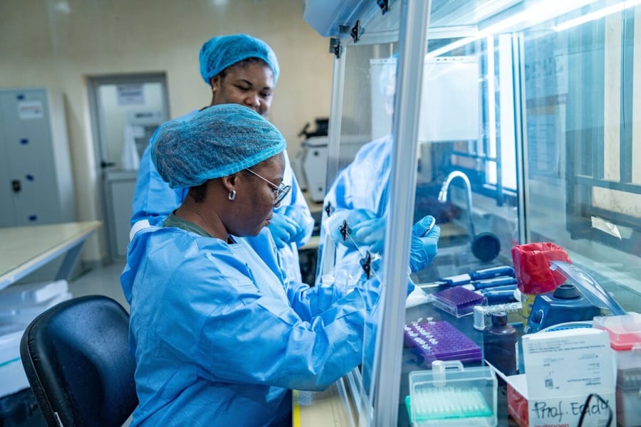 Two laboratory workers in blue gowns and hair coverings handle sample tubes at a safety cabinet in a Congo Ebola testing laboratory