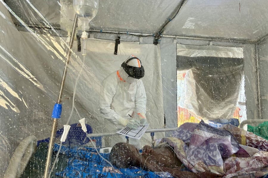 A health worker in a protective suit and face shield writes on a clipboard beside a patient with an IV drip inside a plastic-walled field tent