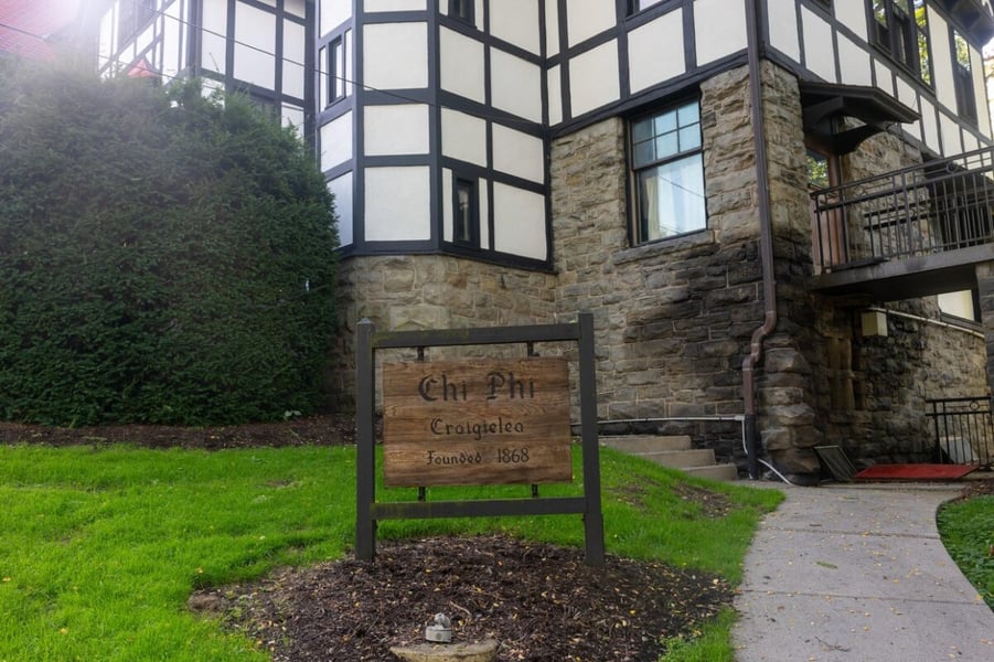 A wooden Chi Phi Craigielea sign on the lawn in front of a half-timbered stone fraternity house in Ithaca