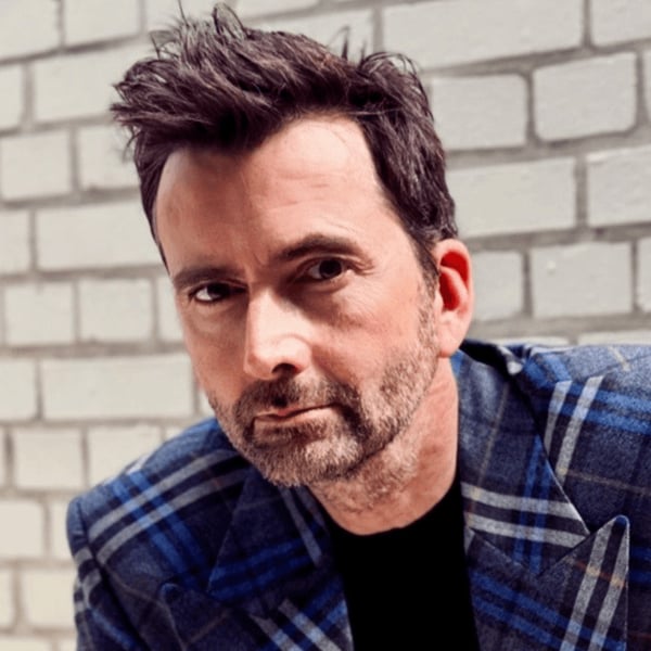 David Tennant in a blue tartan jacket looking at the camera against a white brick wall