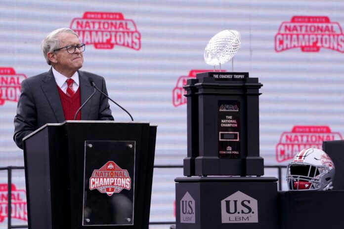 Ohio Gov. Mike DeWine speaks at a podium beside the national championship trophy at an Ohio State rally