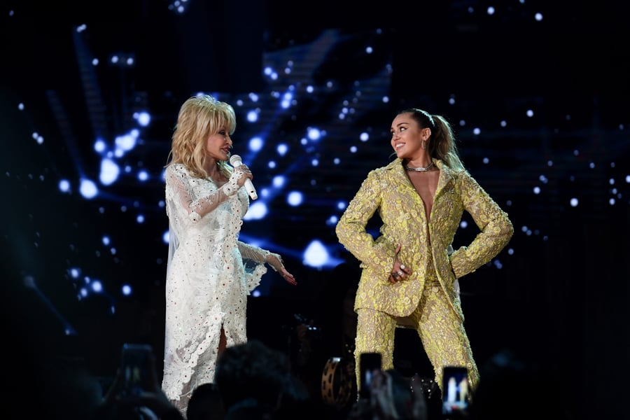 Dolly Parton in a white gown and Miley Cyrus in a yellow suit face each other on stage at the 2019 Grammy Awards