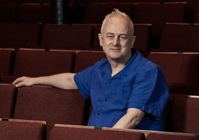 Dominic Cooke in a blue shirt sitting among red theatre seats at the Almeida Theatre