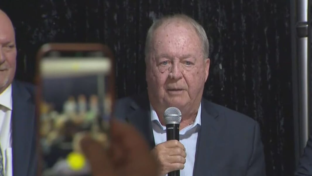 Doug McCallum holding a microphone as he gives a victory speech, with a supporter filming on a phone in the foreground