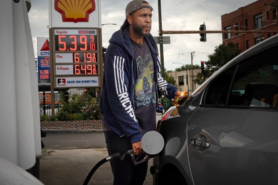 A man in a cap and hooded jacket holds a fuel nozzle at a Shell gas station in Chicago, with regular gasoline priced at $5.39 on the sign behind him