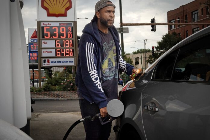 A man in a cap and hooded jacket holds a fuel nozzle at a Shell gas station in Chicago, with regular gasoline priced at $5.39 on the sign behind him