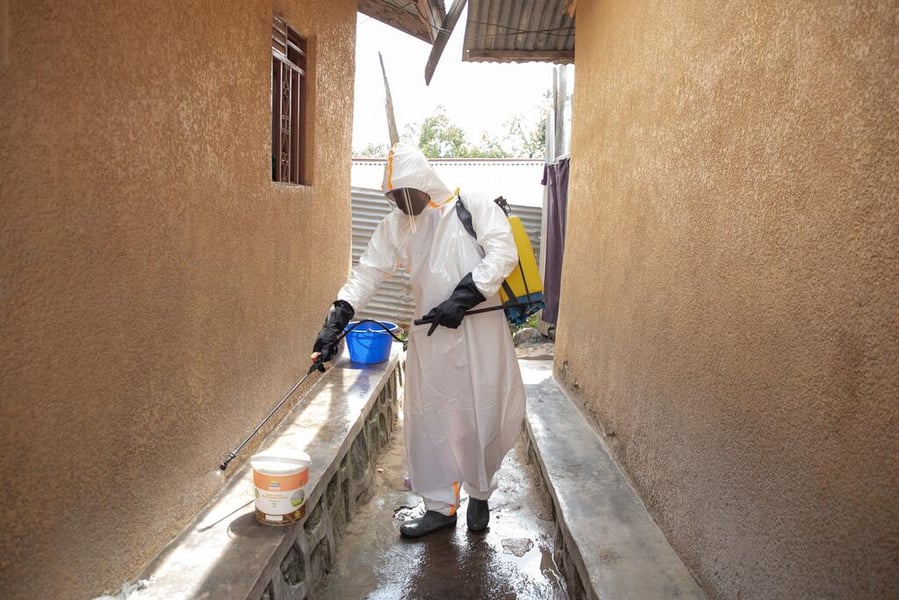 A decontamination worker in a white protective suit and hood sprays disinfectant along a narrow alley between two buildings in Bunia