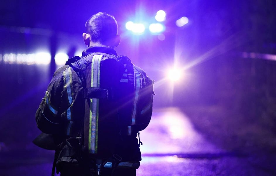 A firefighter in a reflective jacket seen from behind, standing in the glare of emergency vehicle lights at night