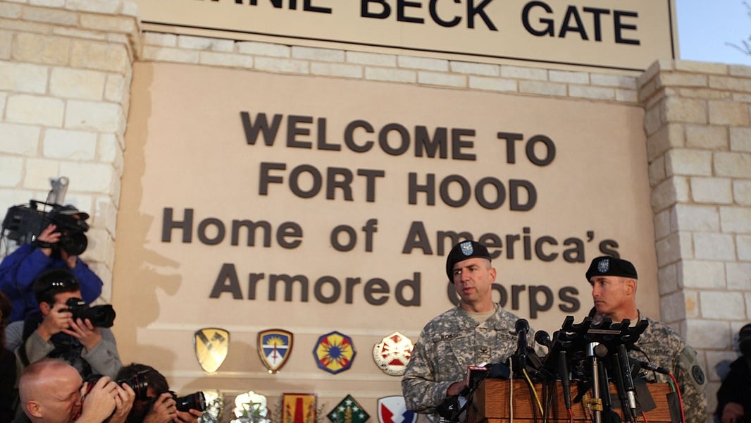 Two Army officers in berets speak at a podium of microphones in front of the Fort Hood entrance sign as photographers look on