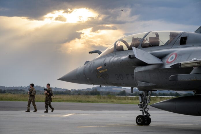 A French Rafale fighter on the apron at Pau-Pyrenees airport under a break in the clouds, with two armed soldiers walking past