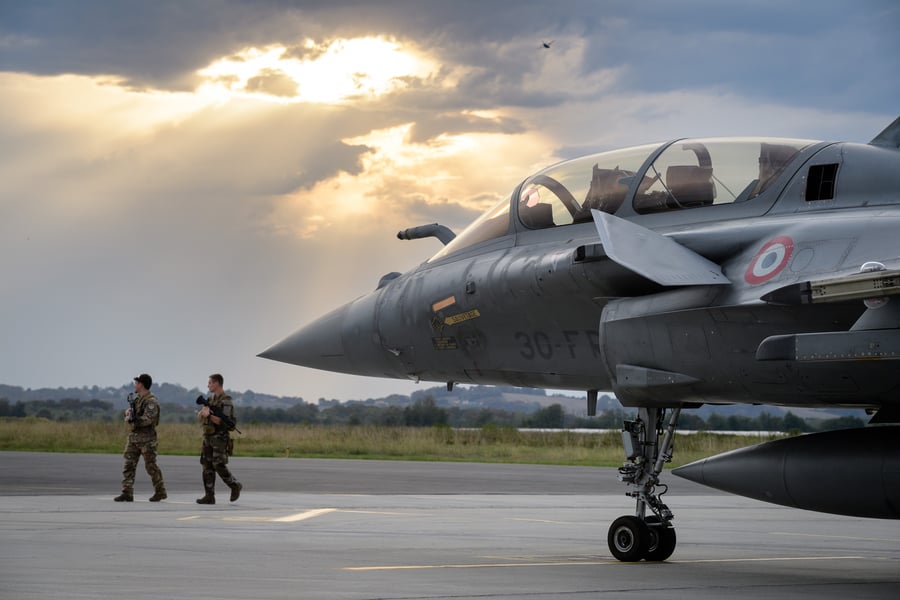 A French Rafale fighter on the apron at Pau-Pyrenees airport under a break in the clouds, with two armed soldiers walking past