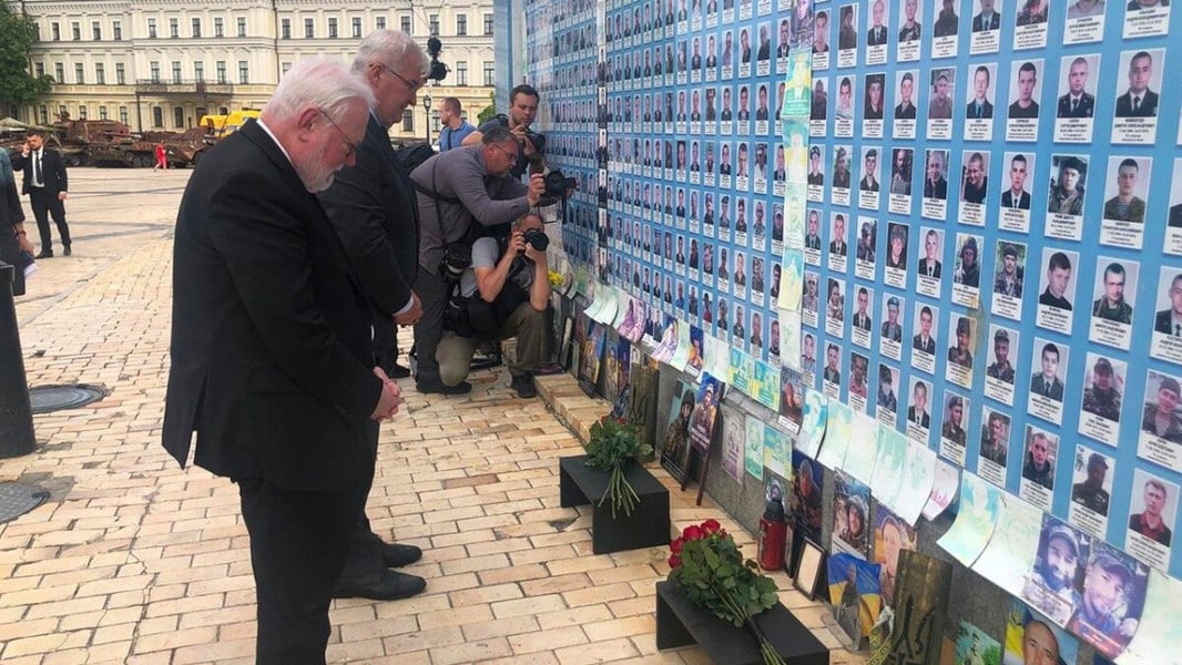 Archbishop Paul Richard Gallagher and a companion bow their heads before the Maidan memorial wall of photographs in Kyiv as photographers kneel nearby