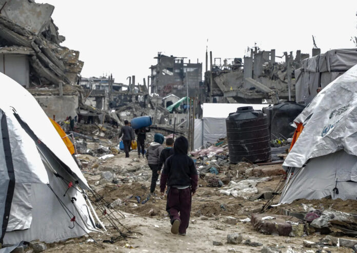 Children walk between tents and rubble in Jabalia camp in the Gaza Strip, past a black water tank and collapsed buildings