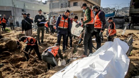 Workers in orange vests dig and stand around a white body bag on churned earth