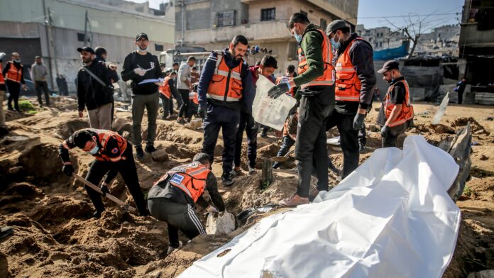 Workers in orange vests dig and stand around a white body bag on churned earth