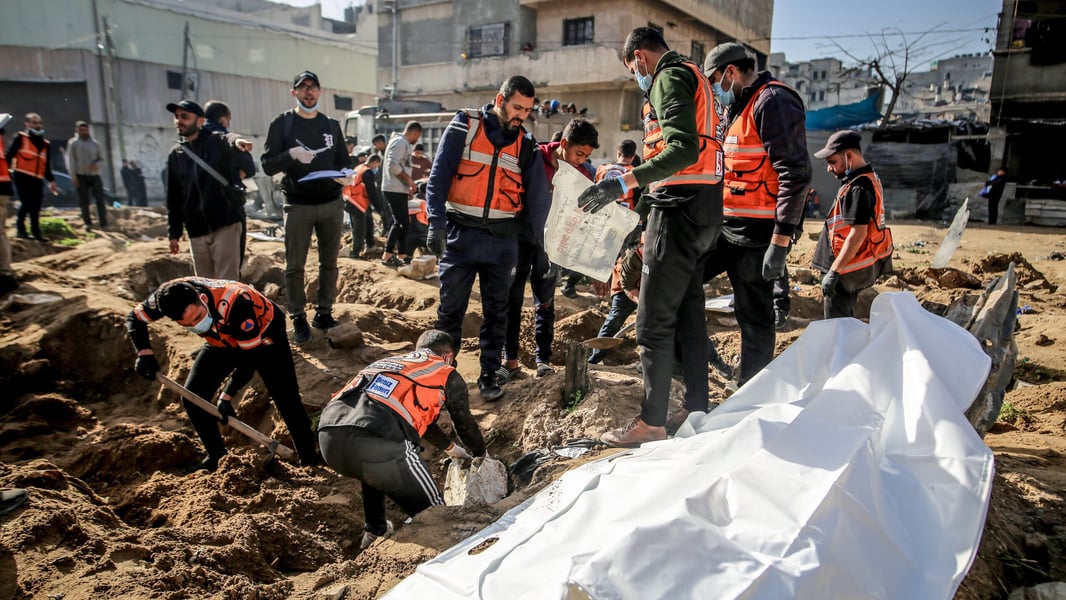 Workers in orange vests dig and stand around a white body bag on churned earth