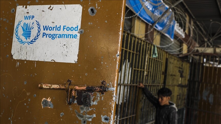 A World Food Programme sign on a weathered door pitted with marks, with a boy standing behind a barred gate