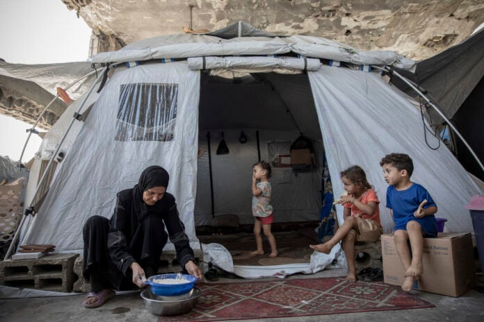 A woman in black sifts flour into a blue bowl outside a grey family tent while three children sit and stand near the entrance