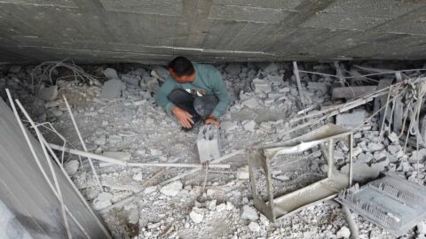 A man crouches among collapsed concrete and twisted metal, salvaging a concrete block from the ruins of a home in Gaza City