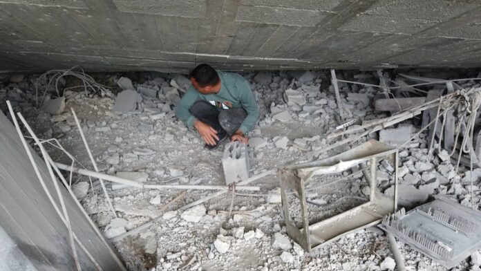 A man crouches among collapsed concrete and twisted metal, salvaging a concrete block from the ruins of a home in Gaza City