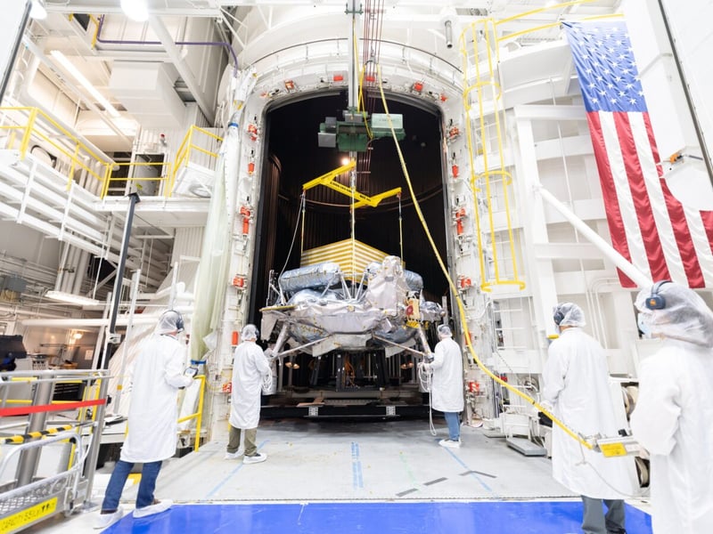 Technicians in white cleanroom suits watch as a crane lowers a lunar lander into a large thermal vacuum chamber at JPL