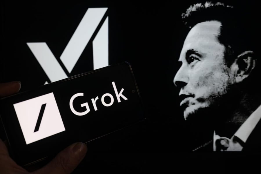 A hand holds a phone showing the Grok logo in front of a screen with the xAI logo and a black-and-white portrait