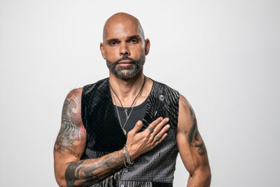 Gus Ormo wearing a black sleeveless top and necklaces, with his right hand on his chest.