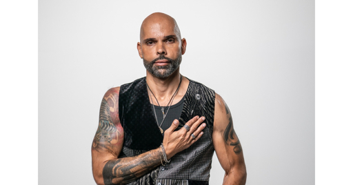 Gus Ormo wearing a black sleeveless top and necklaces, with his right hand on his chest.