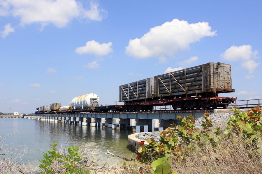 A NASA railroad train with tank cars of helium and other gases crosses a bridge over a Florida waterway under a blue sky
