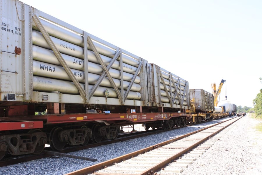 Tank cars stenciled helium sit on a flat car at a railroad yard with a crane and a larger white tank car behind