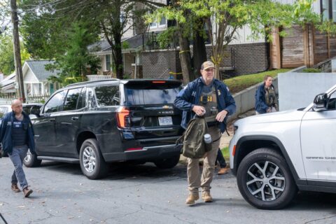 Homeland Security Investigations officers in vests walk past parked SUVs on a residential street