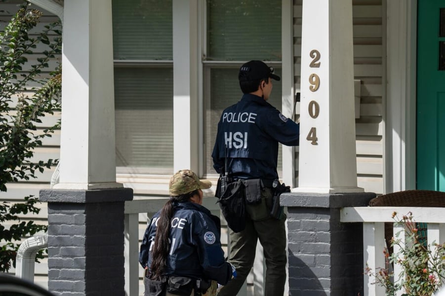 Two officers in jackets marked POLICE HSI stand on a porch during the search