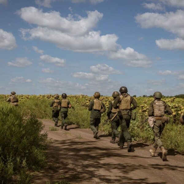 Ukrainian soldiers in body armor and helmets run along a dirt track beside a sunflower field in the Huliaipole district