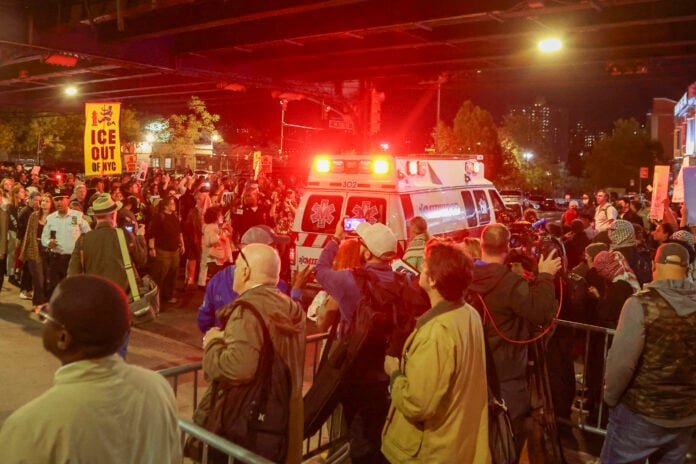 Protesters with an ICE Out of NYC sign crowd a street as an ambulance passes at night after an ICE shooting in New York City