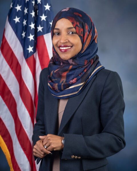 Official portrait of Rep. Ilhan Omar smiling, in a patterned navy headscarf and navy blazer, in front of the US flag