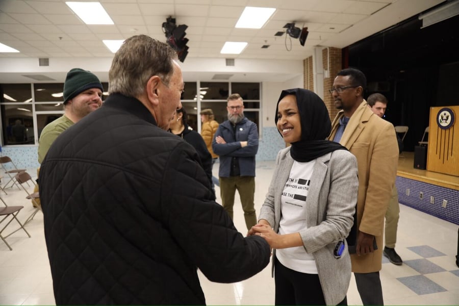 Rep. Ilhan Omar, in a black headscarf and gray blazer, smiles and shakes hands with a man at a town hall in a school gymnasium