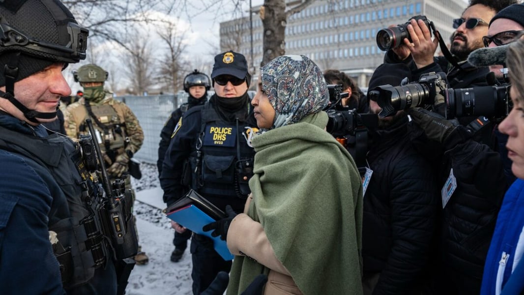 Rep. Ilhan Omar in a green shawl speaking with armed federal officers outside the Bishop Henry Whipple Federal Building in St. Paul as photographers look on