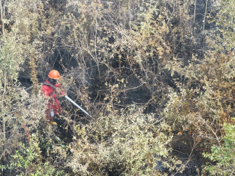 A firefighter in a red suit and orange helmet sprays a hose over blackened ground and dry shrubs, seen from above