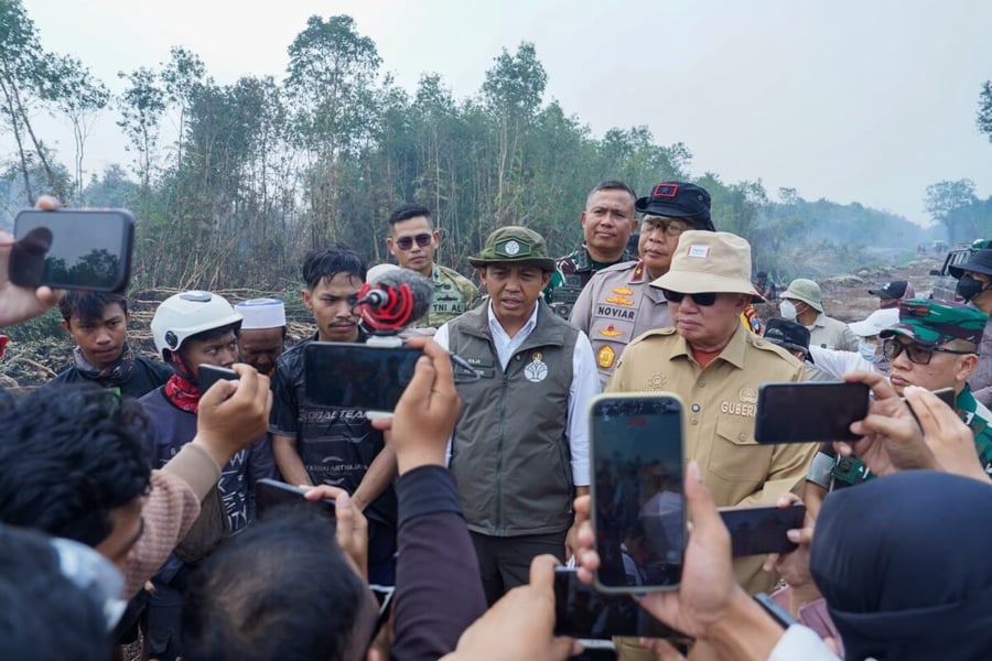 Forestry Minister Raja Juli Antoni speaks to reporters holding phones at a smoky fire site, with a governor and officers behind him